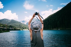 Young man in tan top and panama hat makes photos with digital mirrorless camera of alpine lake with blue water and mountains, nomad explorer, hipster millennial