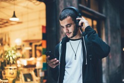 Hispanic young attractive man stands in dark street in front of shop, changes songs and tracks on smartphone, listens to music in wireless headphones. Hipster with slight beard