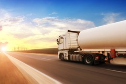 A big metal fuel tanker truck shipping fuel on the countryside road in motion against a blue sky with a sunset