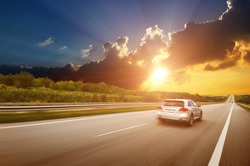 A silver crossover car driving fast on the countryside asphalt road against night sky with clouds and a beautiful sunset