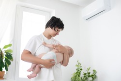 Mother standing picks up baby near the air conditioner