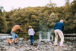 Little boy is skimming pebbles on a lake with his father and grandfather.