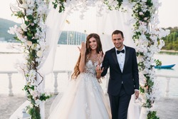 Happy family of groom and bride at wedding day ceremory with arch on background shows their hands with rings. Smiling newlyweds together in wedding suits. Concept of love and happiness