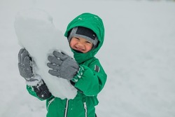A portrait of a funny boy, a child with a large piece of snow in his hands. A kid in a green jumpsuit holds snow in his hands. winter games on the street.