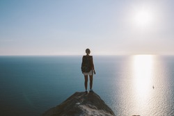 Young woman hiker with backpack standing on cliff and looking forward on the background of the sea, sky. lady tourist on top of a mountain enjoying view. 