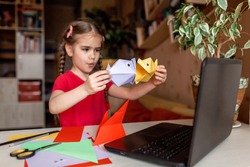 Cute little elementary schoolgirl doing origami fish with folded color paper looking video on laptop, online workshop, kids at-home activity, creativity and distant education, focus on fish