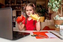 Cute little elementary schoolgirl doing origami fish with folded color paper looking video on laptop, online workshop, kids at-home activity, creativity and distant education