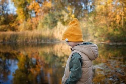 Pensive little boy stands on the bank of river, admires the magnificent view of autumn forest and its reflection in the lake water. Autumn walk. Outdoor lifestyle, active family lifestyle