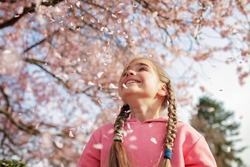 A happy girl in a pink hoodie is throwing sakura petals like those that it's snowing in spring. She rejoices in the coming of spring. Spring vibes. Emotional lifestyle portrait. Selective focus