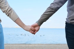 Young man and woman hold hands against the backdrop of blue water and sky. Close-up of hands. Horizontal photo