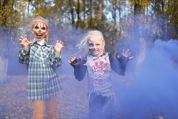 Two children girls girlfriends in Halloween costumes and with makeup make frightening grimaces against the background of autumn leaves and purple smoke. Horizontal photo
