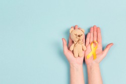 Girl hands holding children's toy with a Childhood Cancer Awareness Yellow Ribbon on blue background. Childhood Cancer Day February, 15.