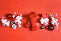 Red dumbbells with winter decorations and gifts on red background. Top view Christmas fitness composition with copy space.  
