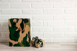 Camouflage shopping bag and gift with khaki ribbon against the white brick wall. Defender of the Fatherland Day, February 23. Space for text.