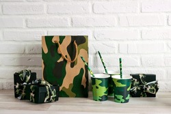 Festive background for Defender of the Fatherland Day, February 23. Party set with camouflage glasses, straws, shopping bag and gifts against the white brick wall. Copy space for text.
