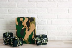 Camouflage shopping bag and gifts with khaki ribbons  against the white brick wall. Defender of the Fatherland Day, February 23. Space for text.