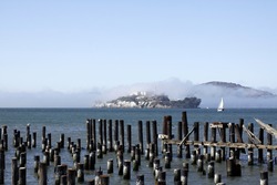 Alcatraz prison fogged over on a sunny day, pilings in the bay in foreground.