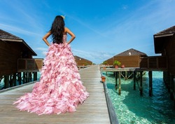 Young bride in beautiful wedding pink dress with elements of feathers. Woman on the background of blue sky and wooden houses.