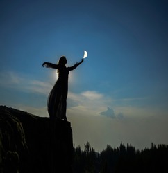 silhoutee of woman standing on top of mountain.  female hold in hand  growing moon above forest trees. backlight and moon light. 