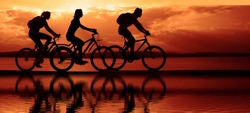 Image of sporty company. friends on bicycles outdoors against sunset sky. Silhouette of motion go of three 3 cyclist along shoreline coast. Reflection sun on water.Copy Space for inscription.threesome