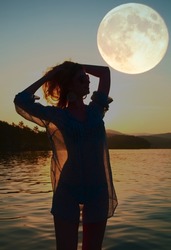 front view unidentified young happy woman stands in warm water. big full moon and calm clear sea water against the backdrop of mountain landscape. clear night sky. Full moon concept