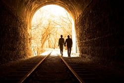 Couple walking hand in hand along the track through a railway tunnel towards the bright light at the other end, they appear as silhouettes against the light. back view.