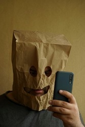 shocked Man with paper packet on head. male looks at a cell phone screen through the eye holes in kraft paper