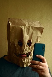 shocked Man with paper packet on head. male looks at a cell phone screen through the eye holes in kraft paper