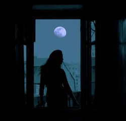 full moon in the night dark  blue  sky. Black silhouette of young slim magical whitch woman looking out of an open balcony window. city, town with many houses on background.