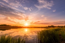 Scenic view of beautiful sunrise or dawn above the pond or lake at spring or early summer morning with cloudy sky background, fog over water and reed grass with dew at foreground. Water reflection.