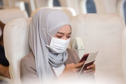 Asian Muslim business woman in hijab headscarf wearing protective face mask and using mobile phone in airplane. Travel and internet connection and coronavirus disease or COVID 19 pandemic outbreak