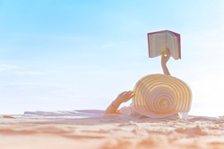 Real view of woman lying on beach, reading book, relaxing on sand. Russian woman in bikini and sun hat, relaxing at sunny, blue sky clear water, remote tropical beaches and countries, travel concept.
