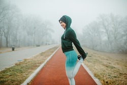 Fit girl is warming up for jogging. Girl is wearing cap, hoodie and tights.