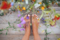 Summer lifestyle portrait of a attractive girl's feet with buds of wildflowers between toes. Staying on wooden terrace surrounded by flowers. Enjoying life, nature. Feet in focus, flowers in blur. 