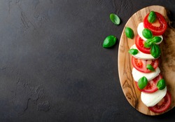 Traditional italian food, sliced tomatoes and mozzarella on a wooden board made of olive tree, dark stone background. Top view, copyspace