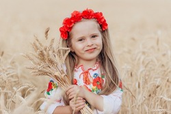 Emotional portrait of a cute and beautiful little girl from Ukraine in a wreath of poppies with blond hair hugging spikelets in a wheat field in the rays of the evening sun. Summer. Harvest concept