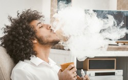 Young man smoking of electronic cigarette at home. Smoker exhales big cloud of smoke, prifile view of arabic guy smokes drinking hot drink while resting at home interior.