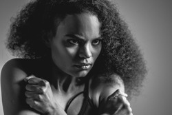 Black and white image. Self-defense, fearful African American girl standing covered with hands in black bare shoulder top isolated on grey background. Human emotions, facial expression concept.