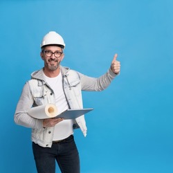 Middle aged handsome architecture man holding roll of wall paper and digital tablet pc in trendy casual denim outfit, hard hat or helmet gesturing thumb up or perfect isolated on blue background. 
