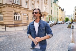 Mature bearded man use walking on urban streets wearing sunglasses and denim jeans shirt. Happy middle aged grey haired business man on old town streets. Travel concept. 