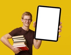 Young student holding a tablet PC with a white blank screen, surrounded by books, studying and using modern technology for academic purposes. Blond man smiles with PDA isolated on yellow background