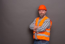 Hipster bearded builder smiling with his arms crossed on dark grey. Builder with orange helmet on ready to work the whole day long.