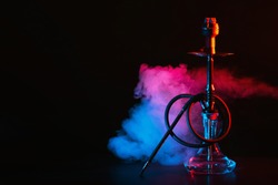 hookah with a glass flask and a metal bowl shisha with colored smoke on the table on a black background with a copy space