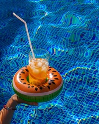 Cold refreshing drink with ice and a straw in the inflatable ring in shape of watermelon in a woman's hand in a pool of blue water. Concept of summer holiday in hotel or pool party. Copy space