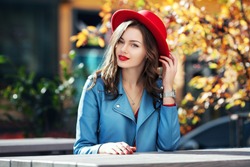 Beautiful happy smiling girl with long hair, red lips, wearing stylish hat, blue jacket posing in autumn street. Outdoor portrait, day light. Female autumn fashion concept. Copy, empty space for text