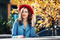 Beautiful happy smiling girl with long hair, red lips, wearing stylish hat, blue jacket posing in autumn street. Outdoor portrait, day light. Female autumn fashion concept. Copy, empty space for text