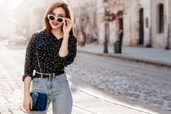 Outdoor portrait of yong beautiful happy smiling woman wearing stylish sunglasses, black polka dot blouse, blue mom jeans, with small quilted bag. Model posin in street of european city. Copy space