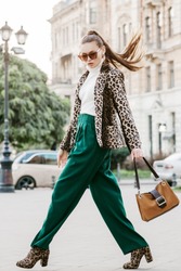 Outdoor full body fashion portrait of  fashionable woman wearing sunglasses, white turtleneck, leopard print blazer, boots, green trousers, holding brown suede bag, walking in street of european city