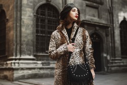 Outdoor fashion portrait of young woman wearing trendy leopard print coat, leather beret, holding black suede bag with fringe, posing in street of European city. Copy, empty space for text 