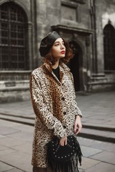 Outdoor fashion portrait of young woman wearing trendy leopard print coat, leather beret, holding black suede bag with fringe, posing in street of European city